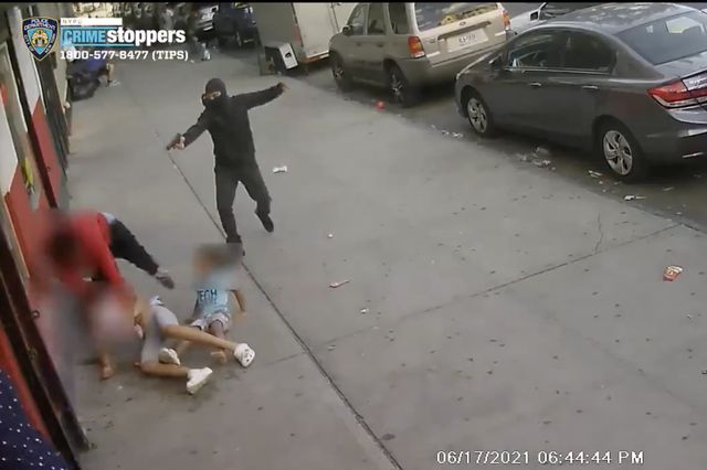 A screen capture image taken from surveillance video showing a man dressed in black aiming a gun at another man dressed in red on the ground as two children are within inches of the shooting.